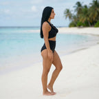 Bikini taille haute gainant noir Elya — silhouette galbée vue de profil sur plage paradisiaque